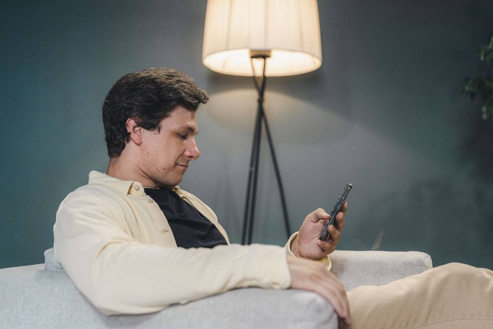 Ein junger Mann sitzt abends auf dem Sofa und schaut auf sein Handy